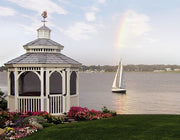 A white gazebo with a gray shingled roof stands on a landscaped lawn beside a body of water, with a sailboat on the water and a faint rainbow in the sky.