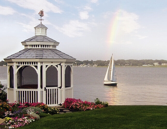 A white gazebo with a gray shingled roof stands on a landscaped lawn beside a body of water, with a sailboat on the water and a faint rainbow in the sky.