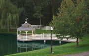 A white arched gazebo bridge with a small octagonal gazebo on a calm pond, surrounded by green grass, trees, and a white fence in a park setting.