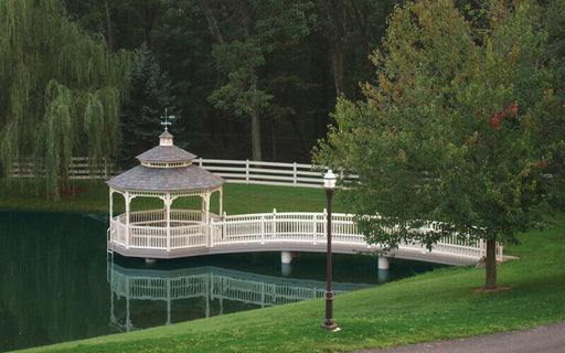 A white arched gazebo bridge with a small octagonal gazebo on a calm pond, surrounded by green grass, trees, and a white fence in a park setting.
