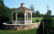 A white gazebo with a brown shingled cupola roof sits on a curved stone retaining wall in a landscaped garden or park setting with green grass and trees under a blue sky.