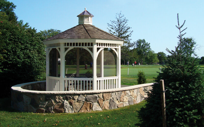 A white gazebo with a brown shingled cupola roof sits on a curved stone retaining wall in a landscaped garden or park setting with green grass and trees under a blue sky.