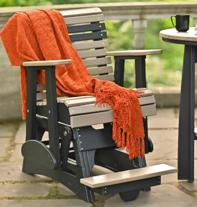 A black and light beige outdoor glider chair with a bright orange throw blanket draped over it, set on a stone patio next to a small table with a black mug.