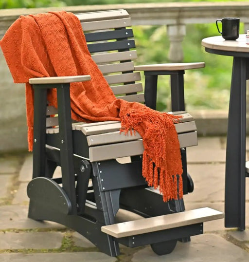 A black and light beige outdoor glider chair with a bright orange throw blanket draped over it, set on a stone patio next to a small table with a black mug.