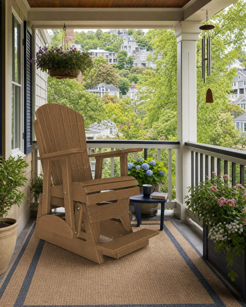 A light brown Adirondack-style glider chair sits on a woven porch rug on a covered front porch, with a small blue side table, potted plants, and a view of a green hillside neighborhood in the background.
