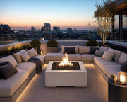 32-inch Alea concrete and limestone fire pit with black lava rocks surrounded by sectional seating on a roof top deck with city views.  The seating area is surrounded by potted plants.   