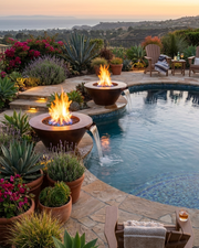Two round outdoor fire pits with glowing flames are arranged beside a stone pool with waterfall spouts, surrounded by potted plants and lounge chairs on a landscaped terrace at sunset.