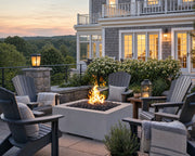 The Alea 32-inch limestone and concrete fire pit on a stone patio surrounded by Adirondack chairs, soft lighting and an east coast home.  