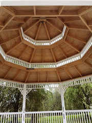 An octagonal wooden gazebo or pavilion viewed from inside, showing a warm brown wood ceiling with white railings and columns, and green trees in the background.