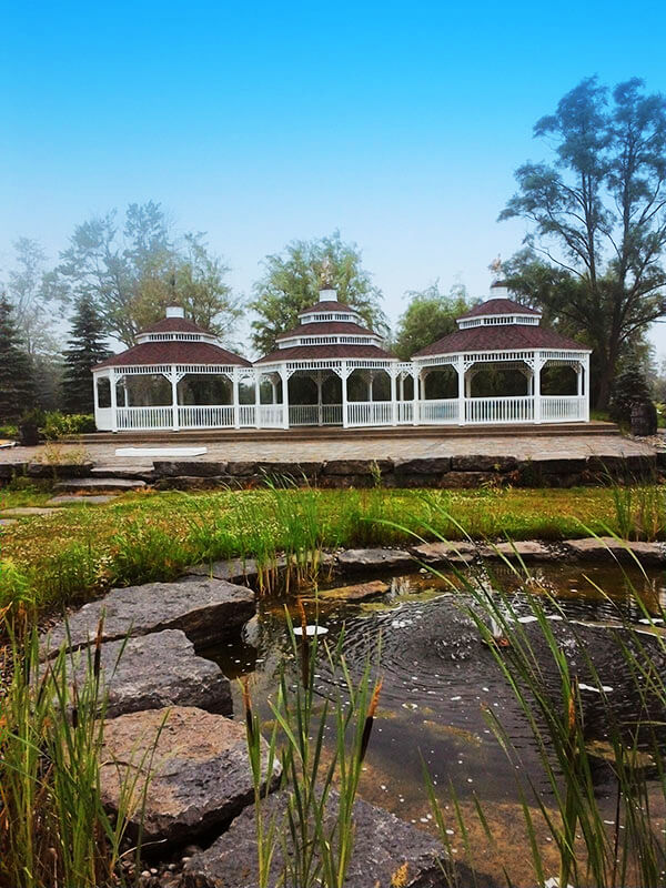 A scenic photograph of a white three-story gazebo with red roofs situated in a landscaped garden with a stone pond, reeds, and trees under a clear blue sky.