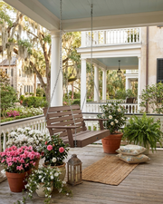 A porch swing hanging from chains on a spacious porch with white columns, surrounded by potted pink and white flowers, green plants, a woven rug, and decorative pillows.