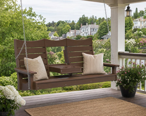 A brown three-person porch swing hanging on a covered deck, with two cream pillows, a small center table, and a garden view in the background.