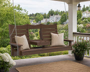 A brown three-person porch swing hanging on a covered deck, with two cream pillows, a small center table, and a garden view in the background.