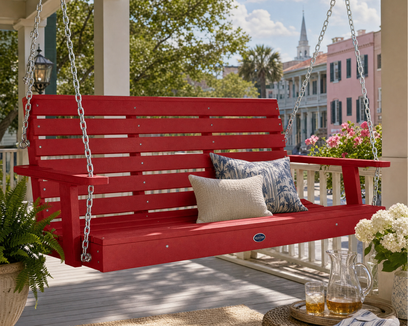 A red  porch swing hanging on a porch with silver chains, styled with two decorative pillows. The porch has a light gray deck, potted plants, and a view of a colorful historic town street in the background.