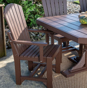 A dark brown Adirondack-style outdoor chair next to a matching rectangular patio table on a woven rug, with a garden and stone border in the background.