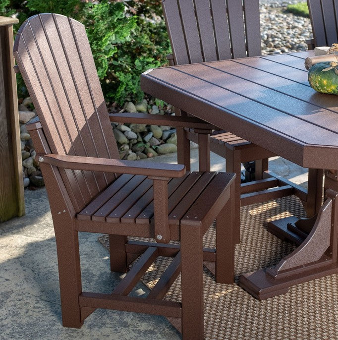 A dark brown Adirondack-style outdoor chair next to a matching rectangular patio table on a woven rug, with a garden and stone border in the background.