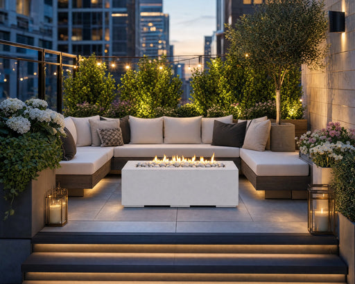 The Alvera 48-inch rectangular fire pit in natural concrete with ivory beach stones centered in front of a sectional outdoor sofa on a terrace with views of the city.  