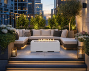 The Alvera 48-inch rectangular fire pit in natural concrete with ivory beach stones centered in front of a sectional outdoor sofa on a terrace with views of the city.  