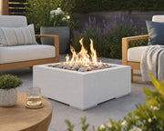 HPC Alea 32-inch concrete fire pit with ivory beach stones and subtle flame on a stone patio centered amongst teak seating, potted plants and flowers. 