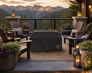 Alea 40-inch-tall vinyl fire pit cover that is covering a fire pit on a stone patio in the mountains.  The fire pit is centered amongst Adirondack chairs, pillows and blankets surrounded by potted plants, flowers and views of the mountains.  