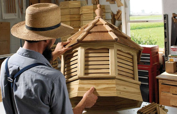 A man wearing a straw hat and blue overalls adjusts a large, handcrafted wooden octagonal gazebo or pavilion structure inside a woodworking workshop.