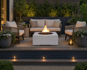 Bravo lightweight natural concrete fire pit with black lava rocks on an outdoor wooden patio.  The fire pit is centered amongst a small seating area that is surrounded by potted plants and flowers.  