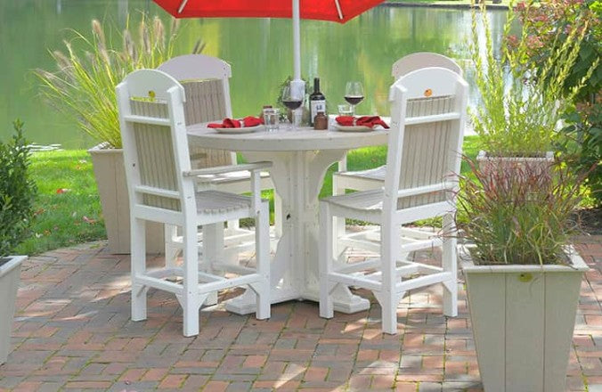 A white outdoor dining set consisting of a round pedestal table and four matching high-back chairs, shown on a brick patio with a red umbrella, plants, and a lake in the background.