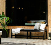 A black outdoor bench with two white throw pillows and a white fringed throw blanket, set on a light wood deck in front of a large black-framed window.
