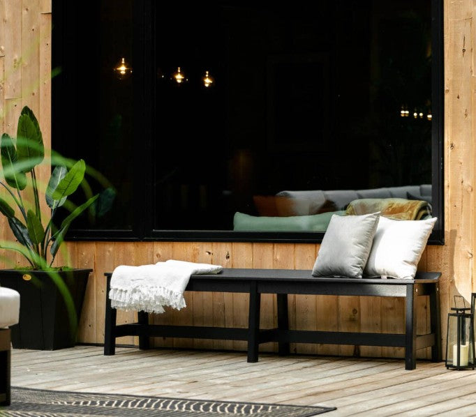 A black outdoor bench with two white throw pillows and a white fringed throw blanket, set on a light wood deck in front of a large black-framed window.