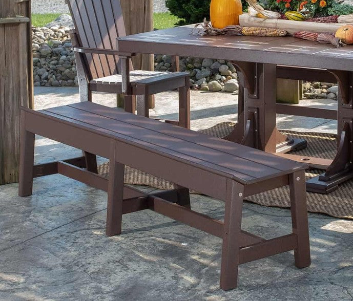 A dark brown outdoor bench positioned in front of a matching wooden dining table on a stone patio, with a wooden chair and fall decorations in the background.