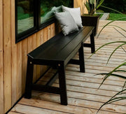 A black bench with two gray throw pillows, positioned on a light wood deck next to a wooden wall and window.