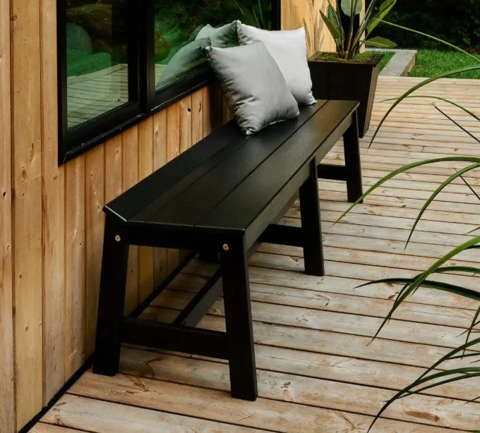 A black bench with two gray throw pillows, positioned on a light wood deck next to a wooden wall and window.