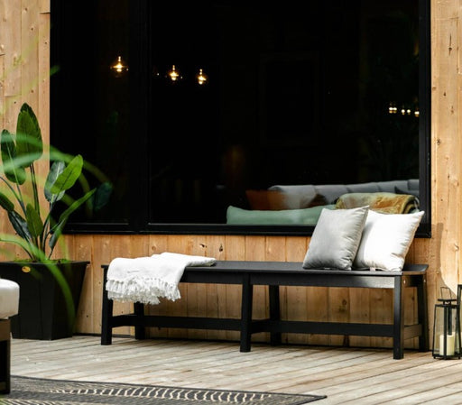 A black outdoor bench with two white throw pillows and a white fringed throw blanket, set on a light wood deck in front of a large black-framed window.