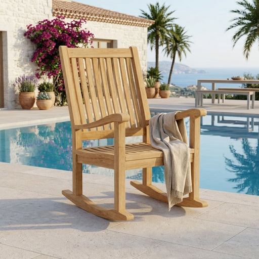 A light natural wood rocking chair with a slatted back and curved arms, placed beside a swimming pool on a patio. A beige throw blanket is draped over the chair, and a white stucco building, palm trees, and ocean are visible in the background.