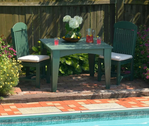 A green outdoor dining set consisting of a rectangular table and two matching chairs with white seat cushions, arranged on a brick patio next to a swimming pool with a wooden fence in the background.