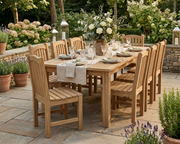 Anderson Teak Chelsea dining table with eight Chelsea dining chairs. The dining set is on a stone patio in a gardn setting and the table is set with white roses in the center.  