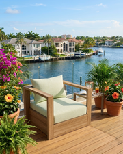 A wooden outdoor armchair with natural  cushions sits on a wooden deck overlooking a waterfront with houses, boats, and potted tropical plants.