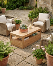 A rectangular light teak coffee table with open shelves sits on a stone patio between two wicker armchairs with cream cushions, surrounded by potted plants and a glass candle holder.