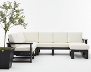 A modern sectional sofa with cream-colored cushions and a black frame, shown in a minimalist studio setting with a potted olive tree on the left.