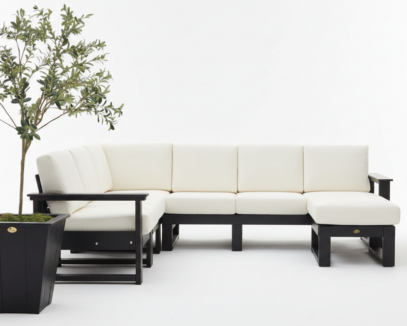 A modern sectional sofa with cream-colored cushions and a black frame, shown in a minimalist studio setting with a potted olive tree on the left.