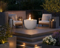 Element natural lightweight concrete firepit with ivory beach stones on an elevated wooden patio with two teak chairs surrounded by potted plants and flowers. 