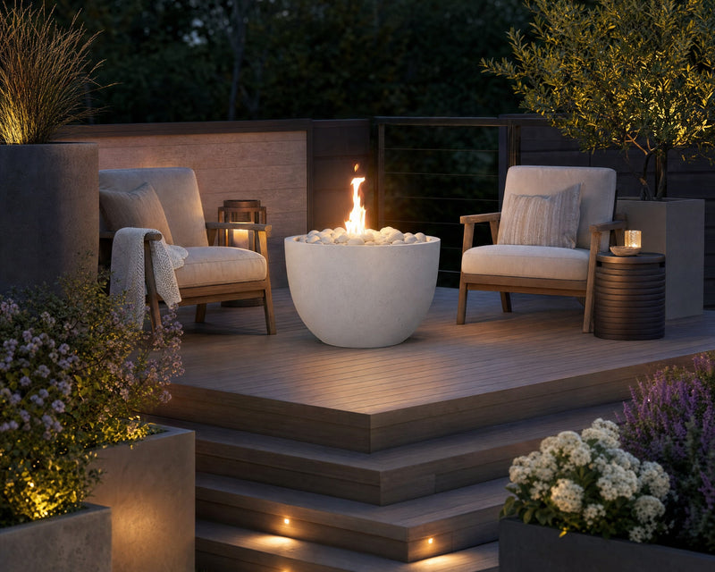 Element natural lightweight concrete firepit with ivory beach stones on an elevated wooden patio with two teak chairs surrounded by potted plants and flowers. 