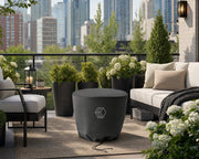 An element fire pit covered with a vinyl cover on a patio in the city.  The patio contains a seating area that is decorated with potted plants and flowers.  