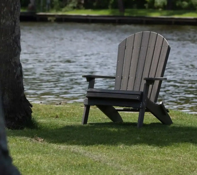 Gray folding Adirondack Chair near a lake on a patch of grass.  