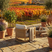 A light wood ARMchair with woven cane details and white cushions sits on a stone patio in a garden setting, with a throw blanket draped over the arm. Terracotta pots, a lemon tree, lavender, and a stone fire pit are nearby, with a vineyard stretching into the background under warm autumn sunlight.