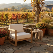 A wooden outdoor patio armchair with thick cream cushions sits on a stone patio beside a small side table with a wine bottle and a throw blanket. A vineyard and hills are visible in the background.