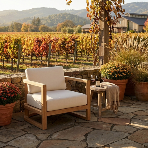 A wooden outdoor patio armchair with thick cream cushions sits on a stone patio beside a small side table with a wine bottle and a throw blanket. A vineyard and hills are visible in the background.