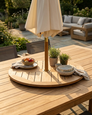 A round wooden outdoor dining table set in a patio garden, featuring a folded beige patio umbrella, bowls, plates, and small potted plants on top.