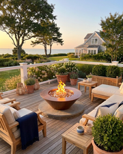Outdoor patio furniture set on a wooden deck overlooking a garden and ocean view at sunset, centered around a round fire pit with cushions, tables, and potted plants.