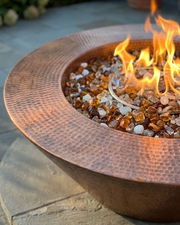 A hammered copper fire pit bowl with a wide rim, filled with amber and clear glass stones and lit with bright orange flames, set outdoors on a stone patio.